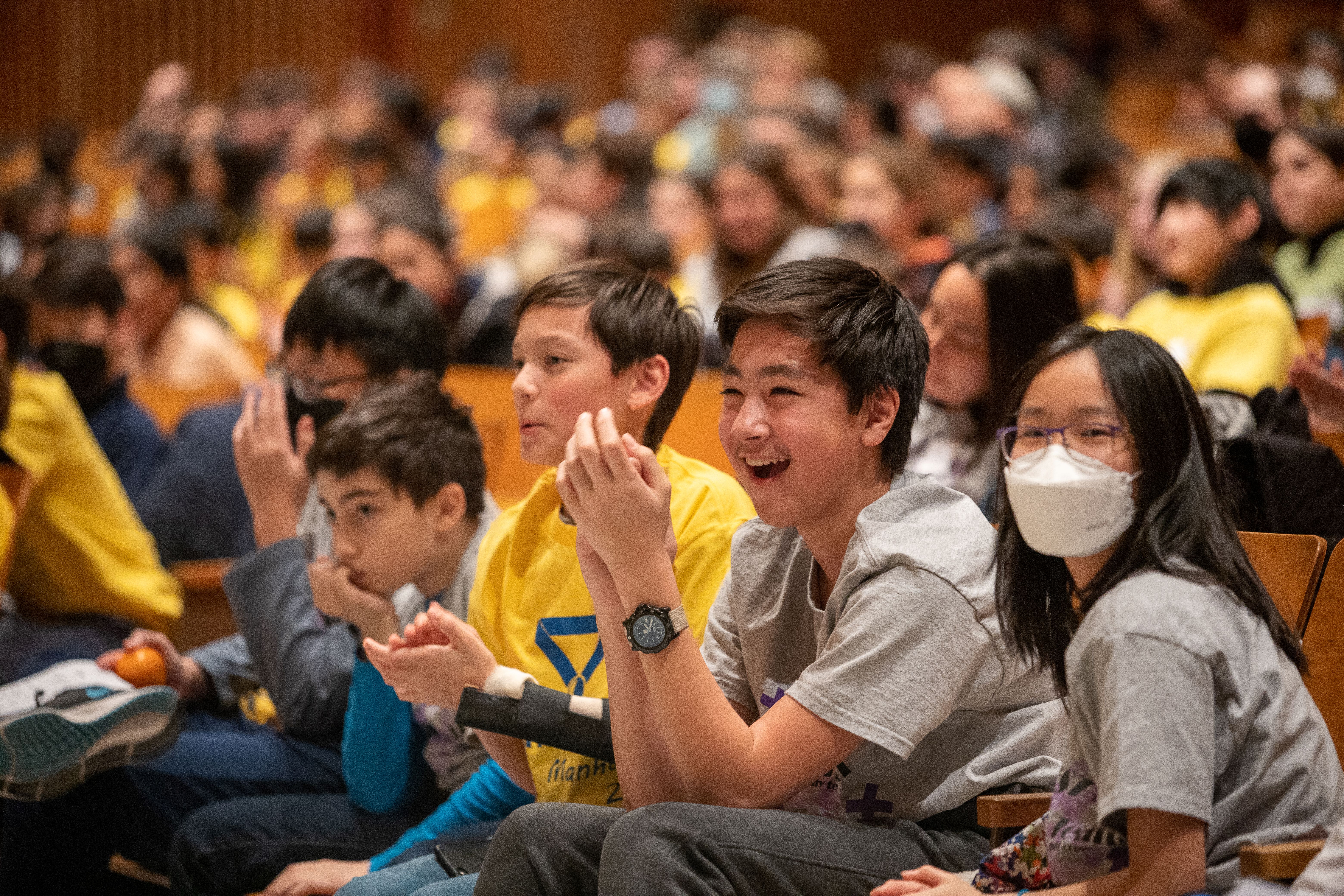 Students smile and clap in an auditorium wearing MATHCOUNTS shirts.